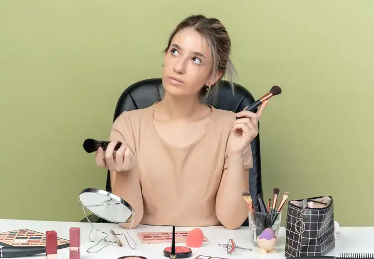impressed young beautiful girl sitting at desk with makeup tools holding makeup brush isolated on olive green background